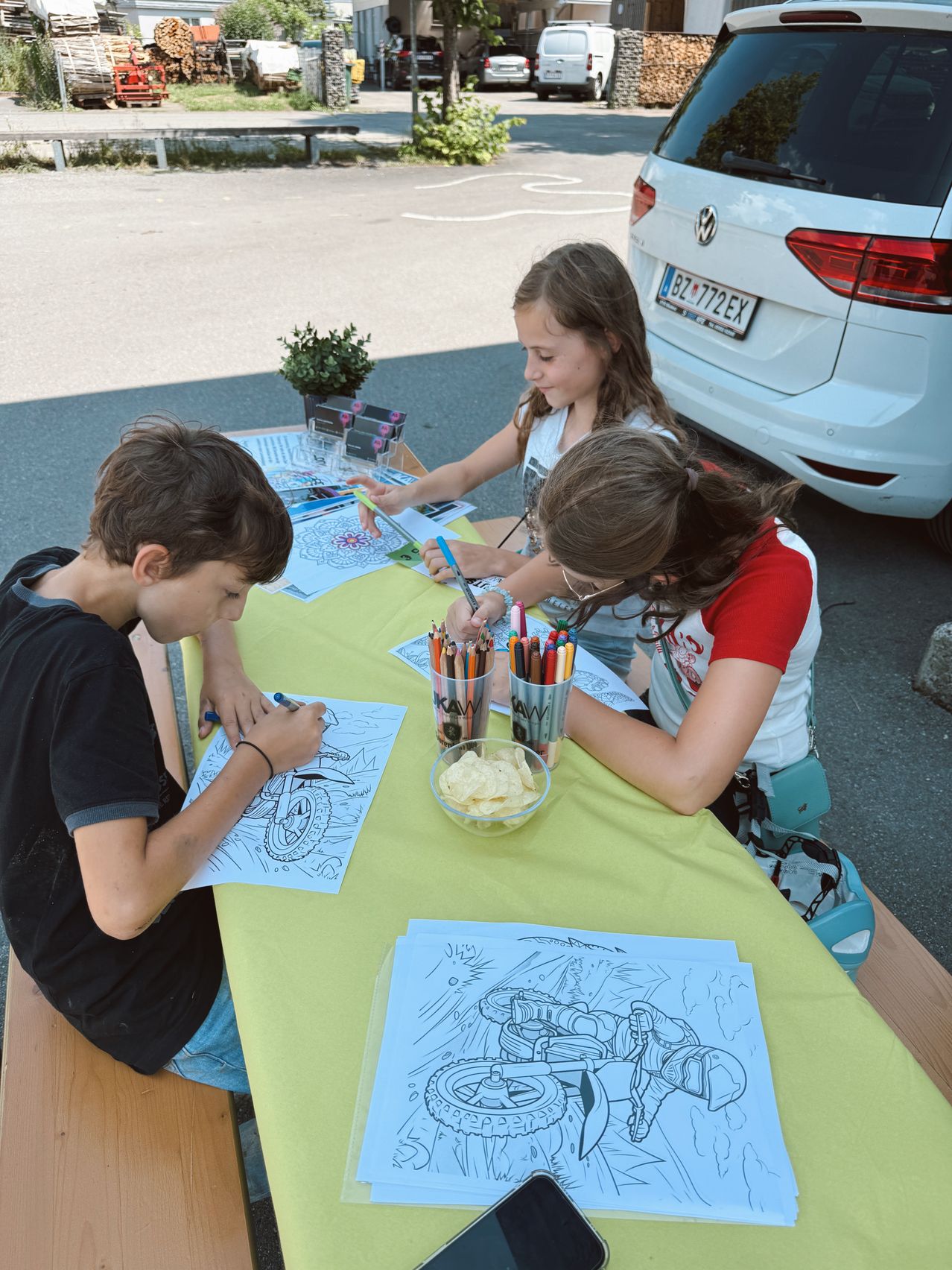 Drei Kinder sitzen um einen Tisch mit Malvorlagen und Buntstiften. Sie sitzen vor einem geparkten weißen Auto mit einem Nummernschild. Eine Schüssel mit Pommes und eine Topfpflanze stehen auf dem Tisch.