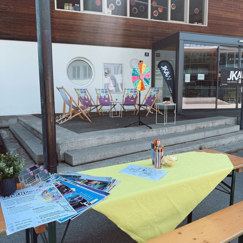 Eine farbenfrohe Einrichtung mit gelber Tischdecke, Stiften und Papieren vor einem Gebäude mit Glastüren. Vier bunte Stühle sind vor einem Drehrad angeordnet. Ein Topf mit Pflanze steht neben dem Tisch.