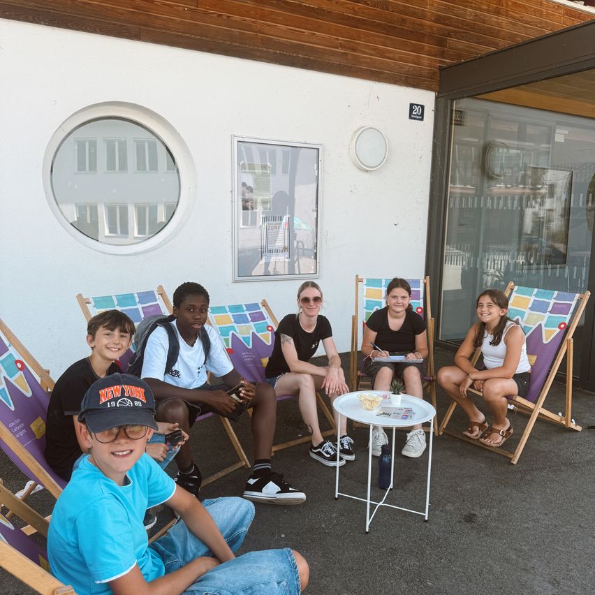 Sechs junge Menschen sitzen auf Klappstühlen im Freien. Einer schreibt, ein anderer hält ein Handy. Ein Tisch mit Snacks und einer Pflanze steht zwischen ihnen. Die Umgebung ist draußen, mit einem Gebäude dahinter.