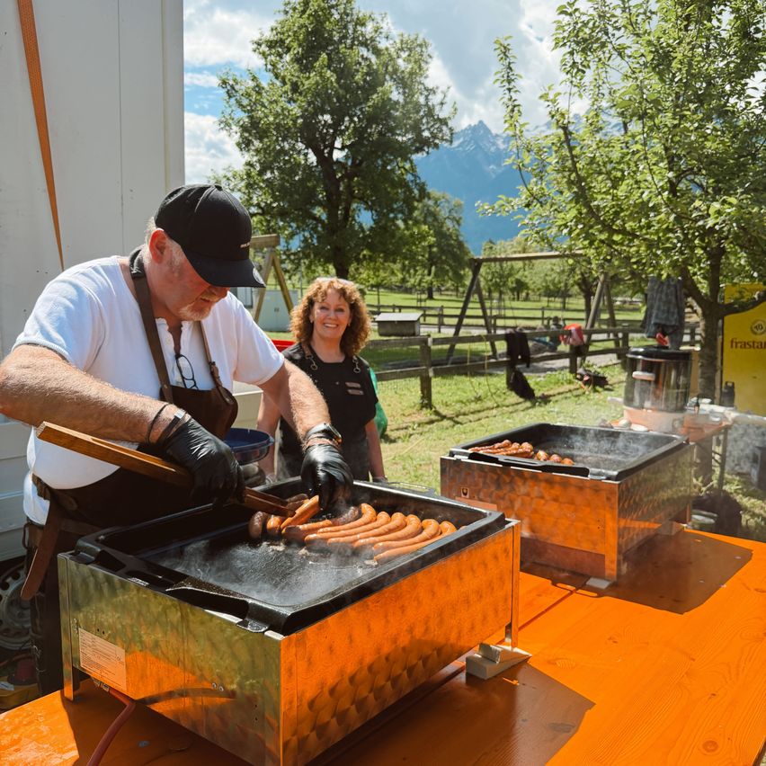 An elderly man cooks sausages on an outdoor grill, with a woman standing nearby. They are surrounded by trees and mountains.