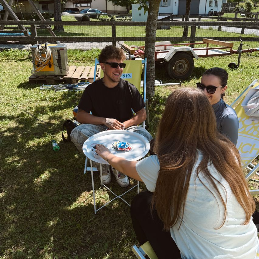 Three people sit in a park with a small table. A man in a black shirt and a woman in a gray shirt sit on chairs, while a woman in a white shirt sits on the grass.