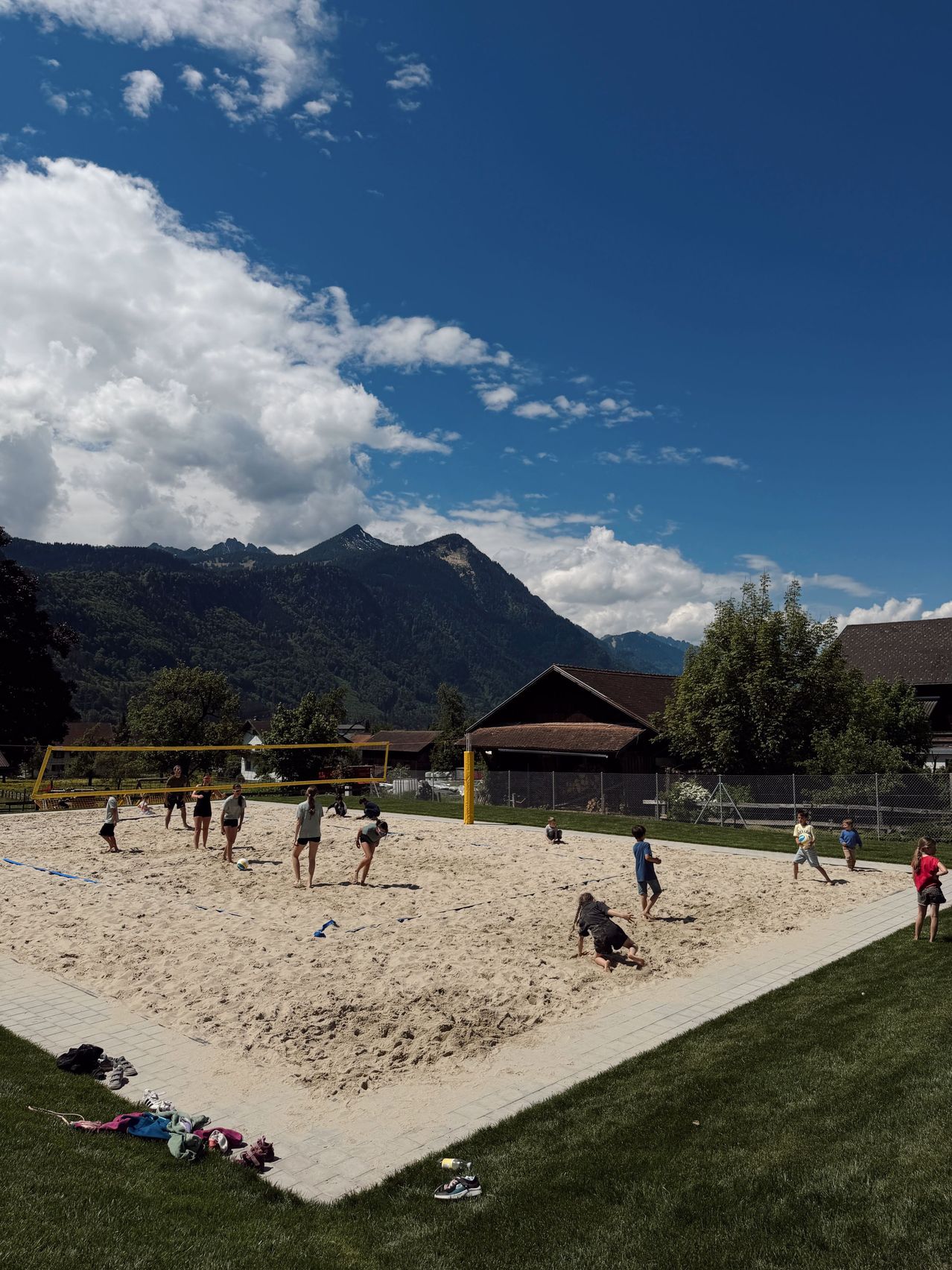 A group of people play volleyball on a sandy court with mountains in the background.