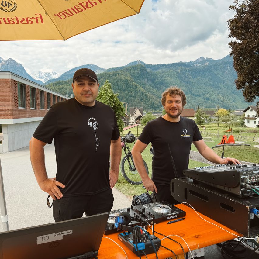 Two men stand behind a table with DJ equipment in front of a building with mountains in the background. One has headphones around his neck, and the other holds his hands on his hips.