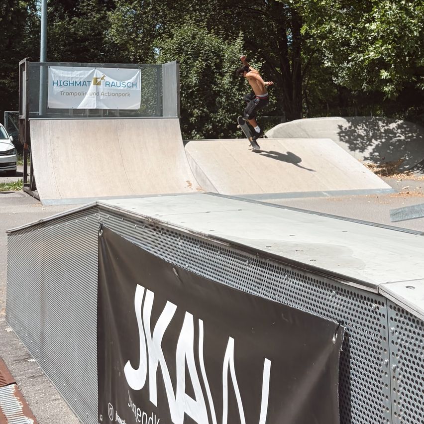 Ein Skateboarder führt eine Trick auf einer Rampe in einem öffentlichen Skatepark aus. Der Park verfügt über Rampen und Banner, darunter eines mit dem Text 'JKAAN'.