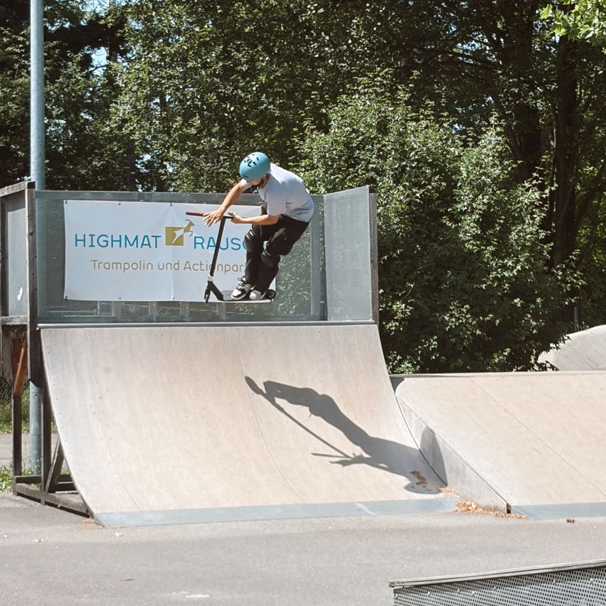 Ein Mann auf einem Roller, mit Helm, führt einen Stunt auf einer Rampe aus. Die Rampe ist von Bäumen und einem Banner mit Text umgeben.
