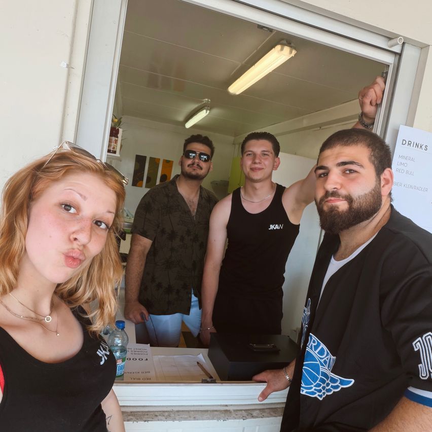 Drei Menschen stehen in einem Raum mit einer Speisekarte an der Wand. Eine Frau posiert für ein Selfie auf der linken Seite, während zwei Männer in die Kamera schauen. Eine Flasche Wasser steht auf der Theke.