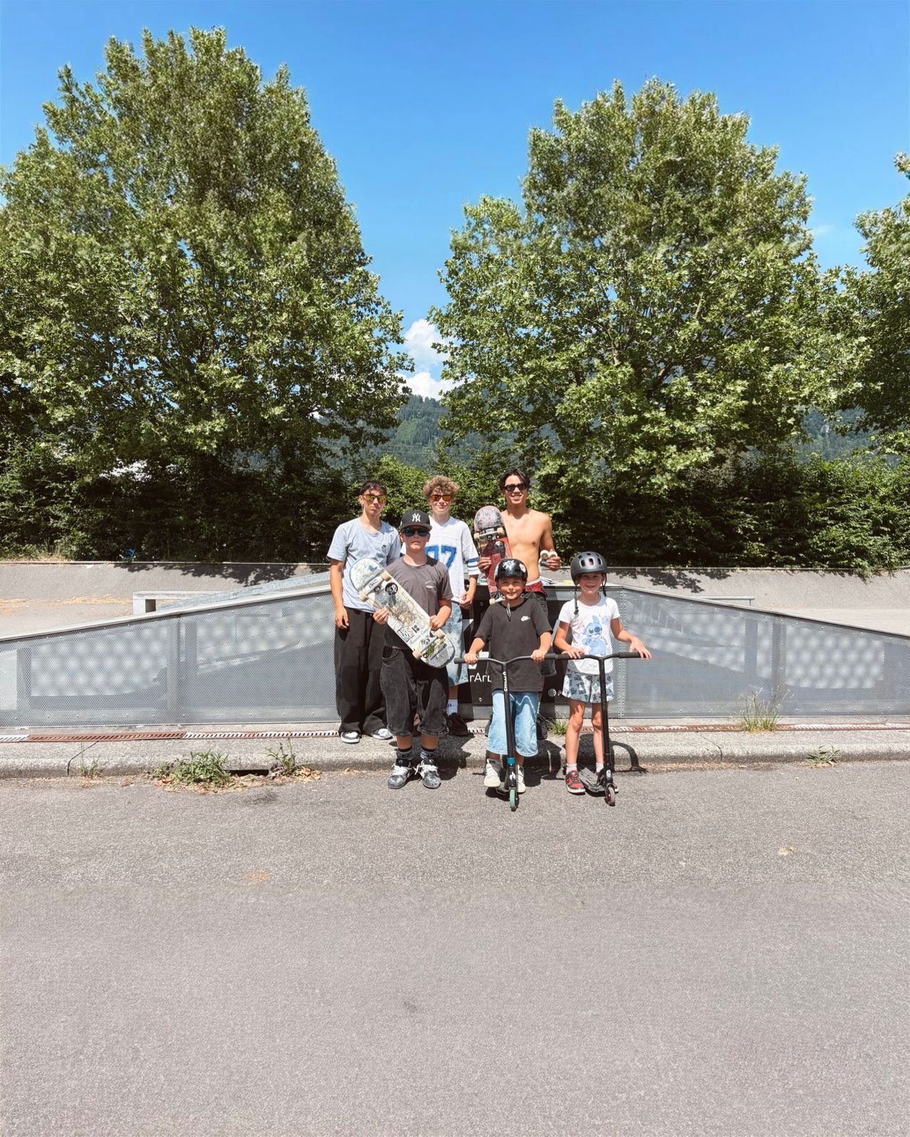 Eine Gruppe von Menschen mit Helmen steht bei einer Skateboard-Rampe, zwei Kinder auf Rollern, eine Person hält ein Skateboard, eine Person ist ohne Oberteil. Bäume und Berge im Hintergrund.