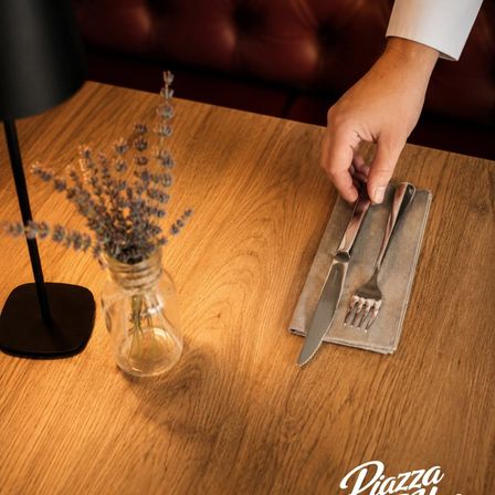 Eine Hand legt ein Messer und eine Gabel auf eine gefaltete Serviette auf einem Holztisch, mit einer Vase mit Lavendelblüten und einer schwarzen Lampe in der Nähe.