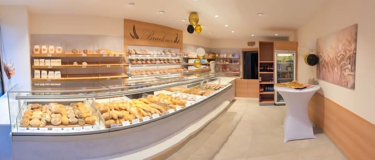 Eine Bäckerei mit modernem Vitrinen-Display voller verschiedener Gebäcksorten. Der Laden hat ein helles Farbschema und die Decke ist mit mehreren runden Lampen ausgestattet.