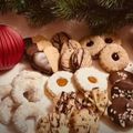 Eine Auswahl an Keksen mit verschiedenen Toppings, Schokoladen und Streuseln auf einer weißen Oberfläche mit einem Weihnachtsbaum im Hintergrund.