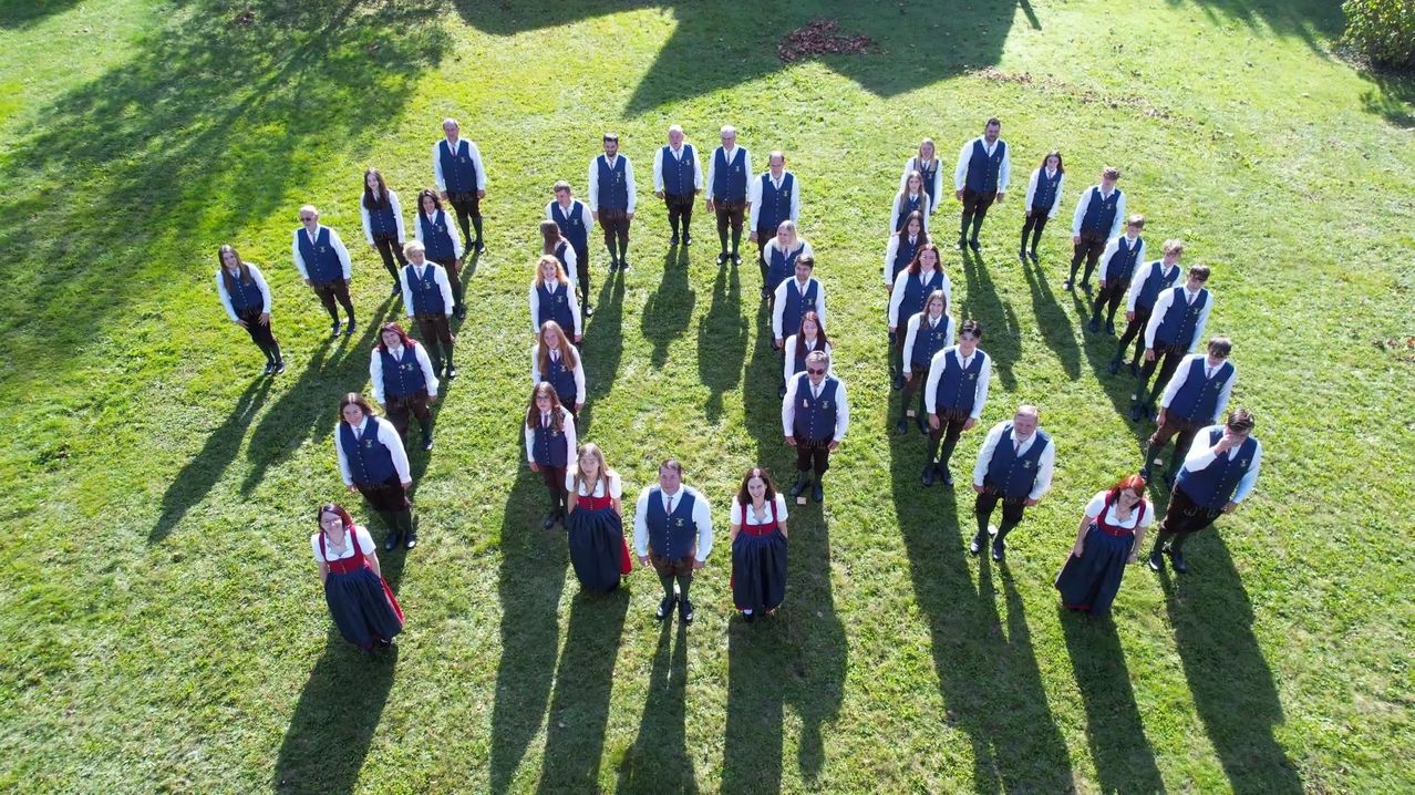 Eine Gruppe von Menschen in traditioneller Kleidung bildet einen Kreis auf einem Rasenfeld, einige tragen Gürtel und Brillen.