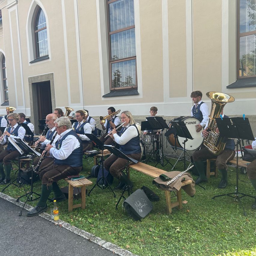 Eine Blaskapelle spielt auf einer Rasenfläche vor einem Gebäude mit Rundbogenfenstern. Die Musiker sitzen auf Holzstühlen und spielen verschiedene Instrumente.