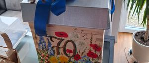 Eine Schachtel mit blauer Schleife und der Zahl 70 darauf, verziert mit Blumen und Bienen, steht auf einem Tisch mit einem Topf.