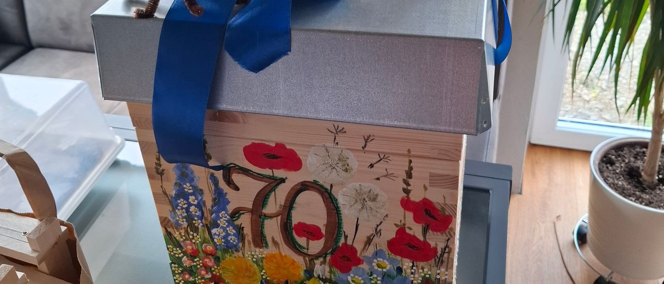 Eine Schachtel mit blauer Schleife und der Zahl 70 darauf, verziert mit Blumen und Bienen, steht auf einem Tisch mit einem Topf.