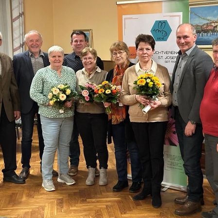 Eine Gruppe von Menschen steht in einem Raum und hält Blumen. Drei Frauen halten im Zentrum Sträuße. Ein Schild im Hintergrund lautet 'Achtverein St. Josef'.