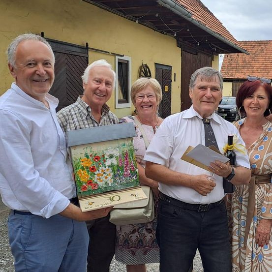 Eine Gruppe von Menschen steht vor einem Haus. Der Mann in der Mitte hält ein Papier und eine Flasche. Der Mann auf der linken Seite hält eine Schachtel. Die Frau auf der rechten Seite trägt ein Kleid und eine Halskette.