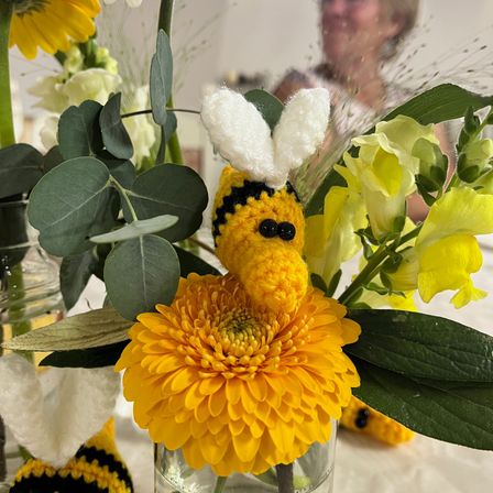 Eine gehäkelte Biene sitzt auf einer gelben Blume, umgeben von grünen Blättern und anderen Blumen. Eine Person ist im Hintergrund leicht zu erkennen.