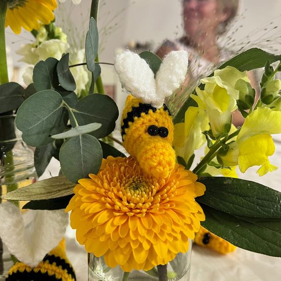 Eine gehäkelte Biene sitzt auf einer gelben Blume, umgeben von grünen Blättern und anderen Blumen. Eine Person ist im Hintergrund leicht zu erkennen.