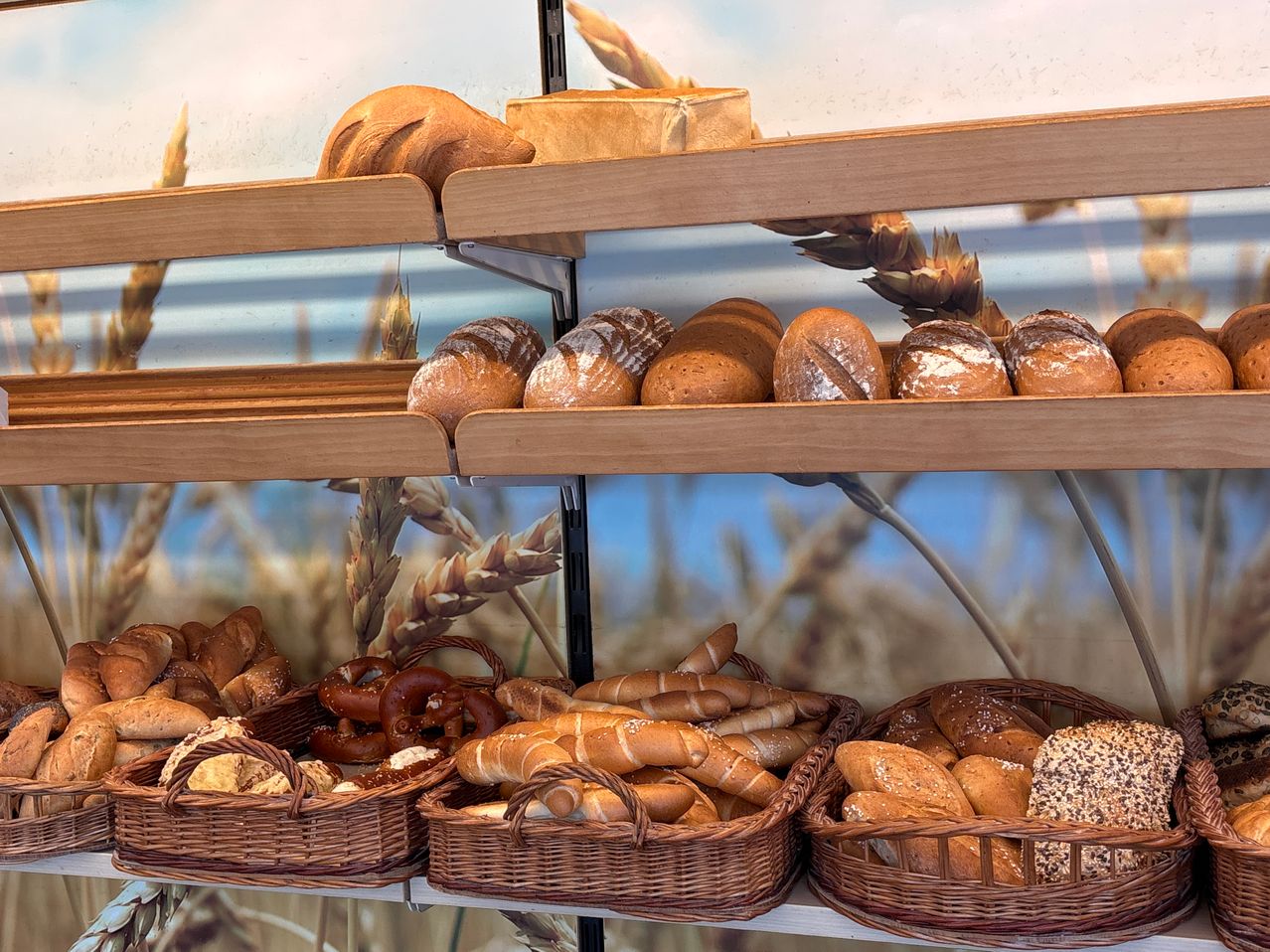 Eine Bäckerei-Auslage mit mehreren Regalen und Körben, gefüllt mit verschiedenen Brotsorten und Gebäck, vor einem Hintergrund aus Weizen.