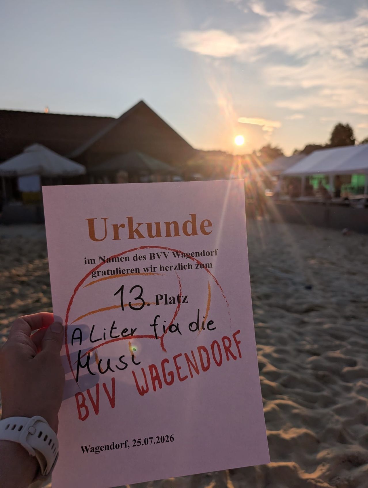 Eine Person hält ein Zertifikat am Strand bei Sonnenuntergang, mit einem Basketballplatz und Zelten im Hintergrund. Das Zertifikat lautet 'Urkunde im Namen des BVV Wagendorf' und '13. Platz A Liter für die Musik BVV Wagendorf'.