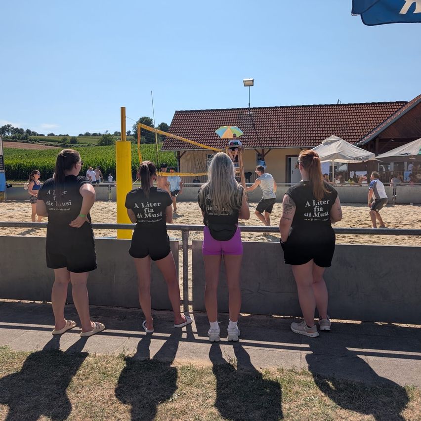Vier Frauen in schwarzen Shirts mit weißem Text beobachten ein Volleyballspiel an einem sonnigen Tag. Sie stehen vor einer Betonbarriere. Im Hintergrund springt eine Person über das Netz.