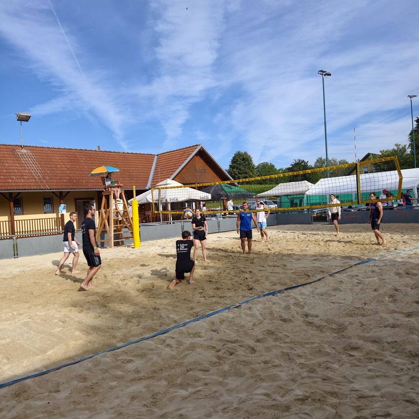 Eine Gruppe von Menschen spielt Volleyball auf einem sandigen Platz mit einem gelben Netz und einem Schiedsrichter auf einer Leiter.