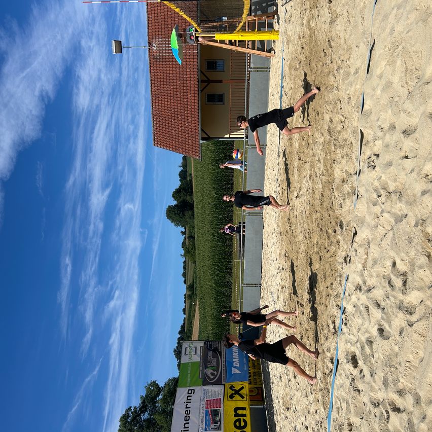 Eine Gruppe von Menschen spielt Beachvolleyball auf einem sandigen Platz mit einer blauen Linie als Begrenzung. Ein Haus mit rotem Dach steht dahinter.