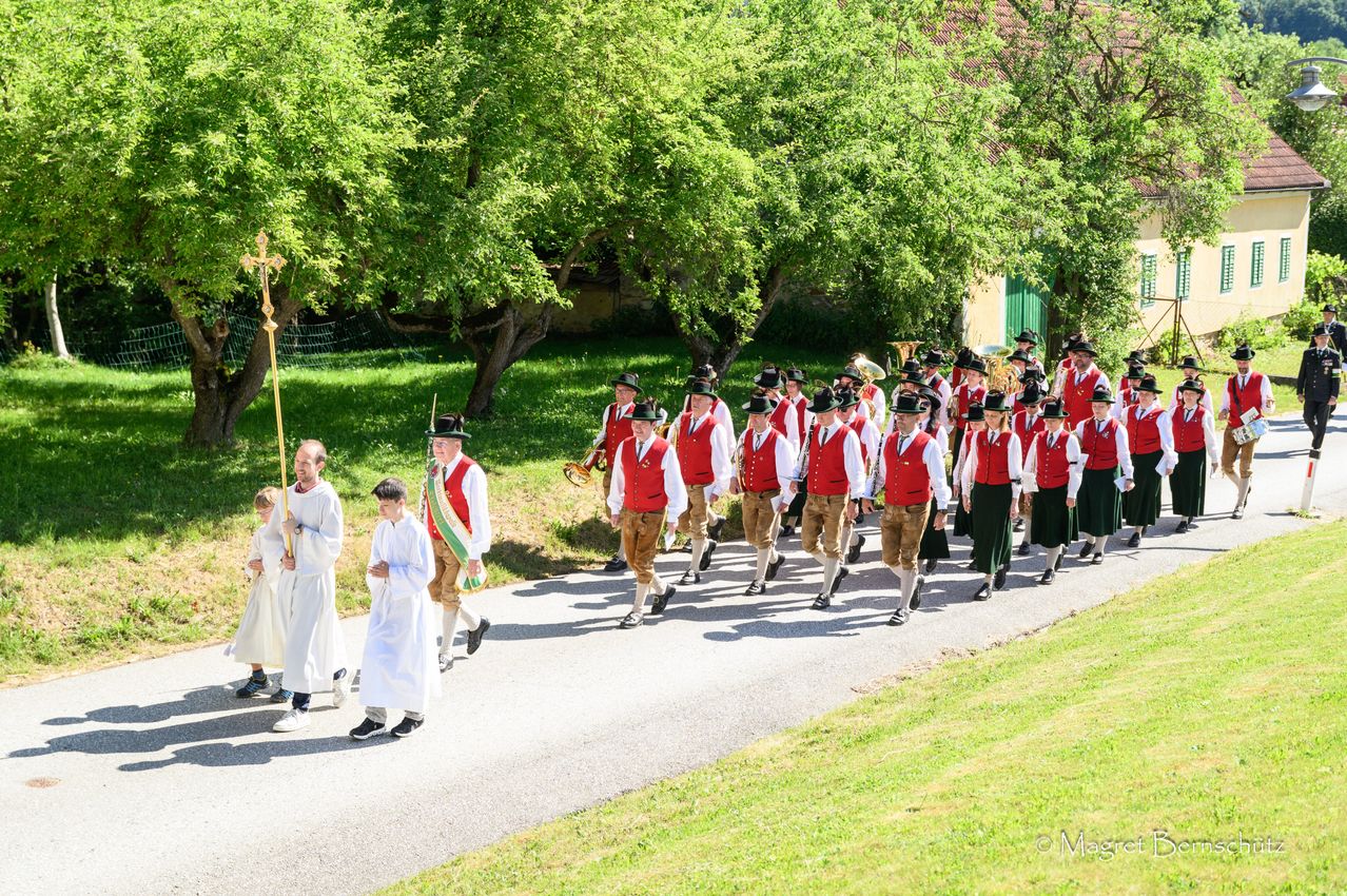 A procession of people in traditional attire is marching on a road, with musicians playing instruments. Two men in white robes lead the procession, followed by others wearing red vests and black hats. Trees and grass are on either side of the road.