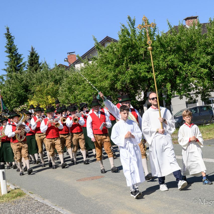 A procession of men in traditional garb playing brass instruments leads two young boys in white robes. A man in sunglasses holds a cross staff, walking beside them. Trees and houses are in the background.