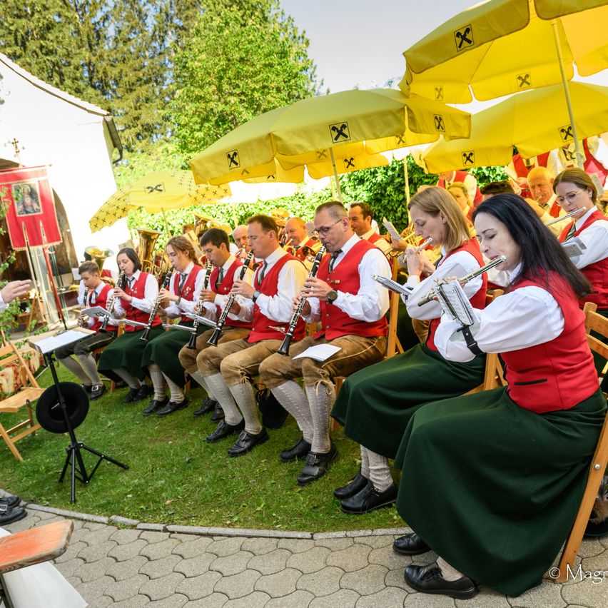 A band in traditional clothing plays instruments under yellow umbrellas, with people sitting on chairs on a lawn. A music stand and a black hat are nearby.