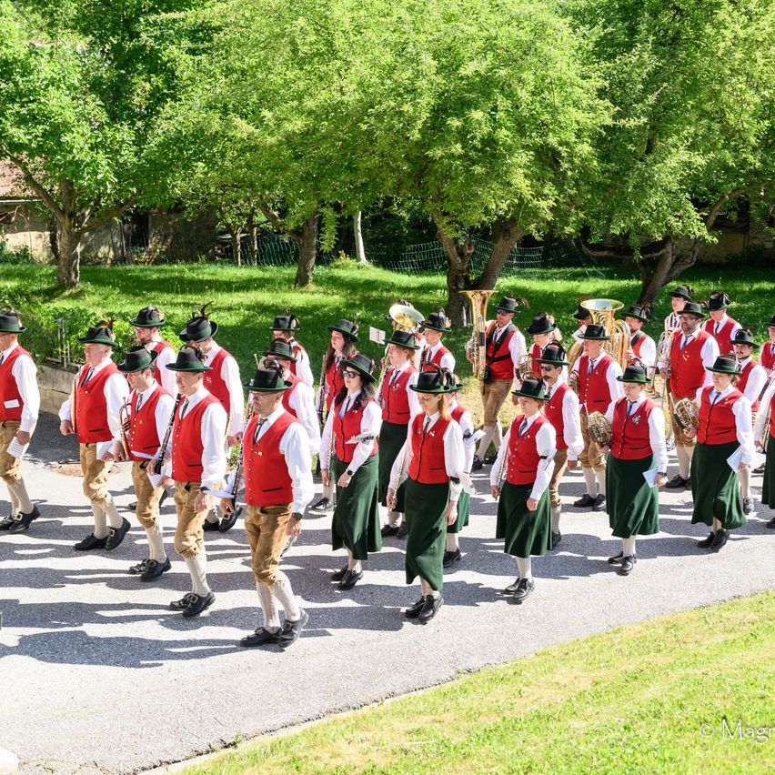 A marching band dressed in traditional red and green attire is walking on the road. The band includes woodwind and brass instruments.