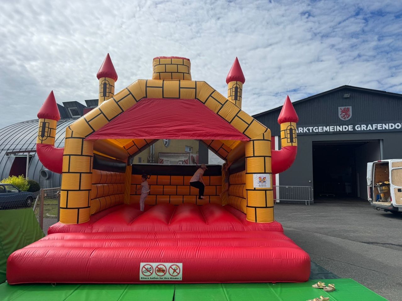 Two children playing in a large inflatable castle on a cloudy day. The castle is red and yellow with a red roof. A sign reads 'No eating and drinking inside.'