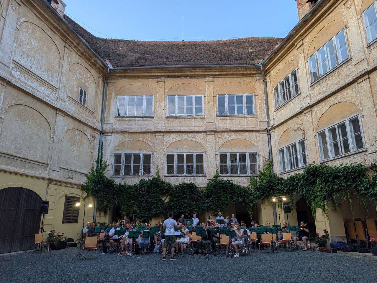 Eine Gruppe von Musikern spielt im Freien, in Stühlen um einen Innenhof herum sitzend, mit einem historischen Gebäude im Hintergrund.