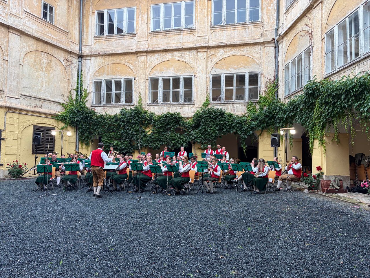 Ein Orchester in traditioneller grüner und roter Kleidung spielt im Freien vor einem alten Gebäude mit großen Rundbogenfenstern und Efeu.