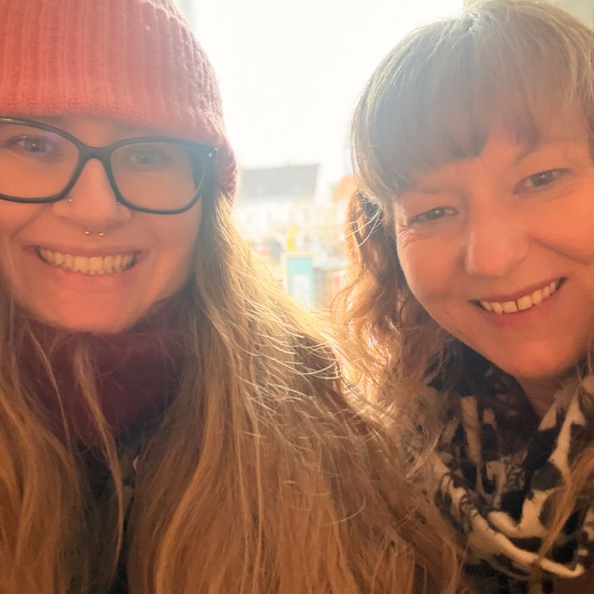 Two women smiling indoors, one with a red beanie and glasses, the other with a scarf. They both have bangs.