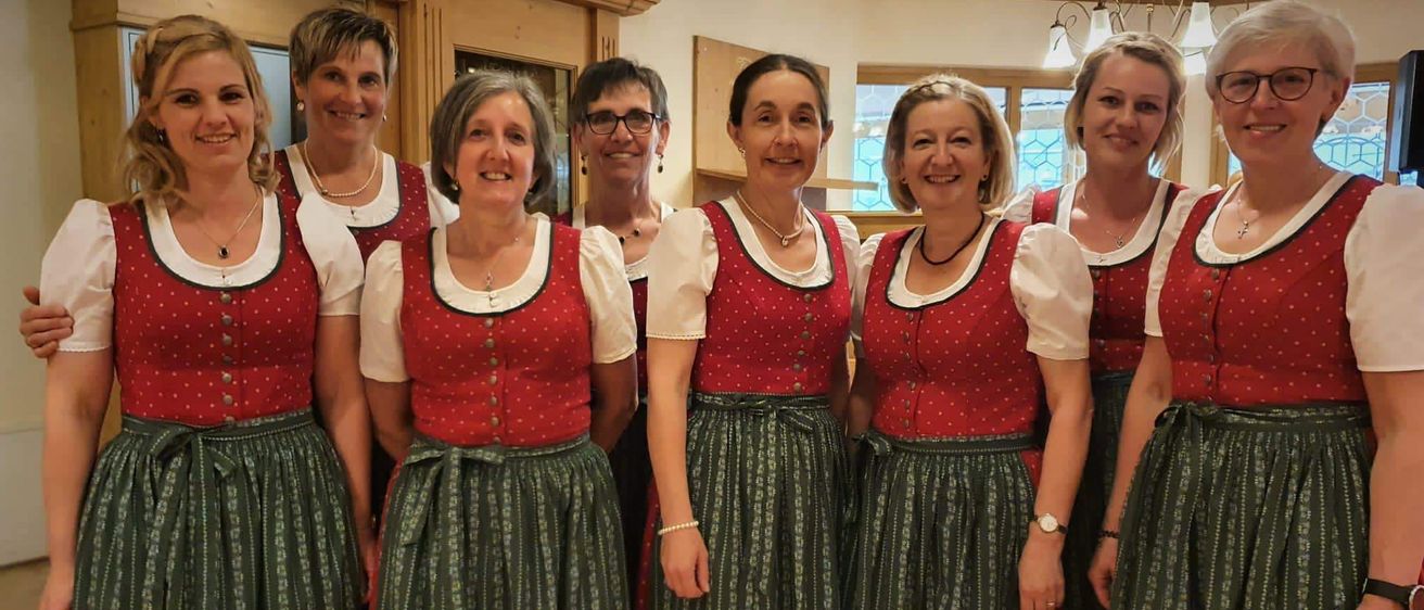 Sechs Frauen in traditioneller bayerischer Kleidung, alle lächelnd, stehen zusammen in einem Raum. Jede Frau trägt eine rote Weste mit weißen Punkten. Sie sind mit Halsketten und Armbändern geschmückt.
