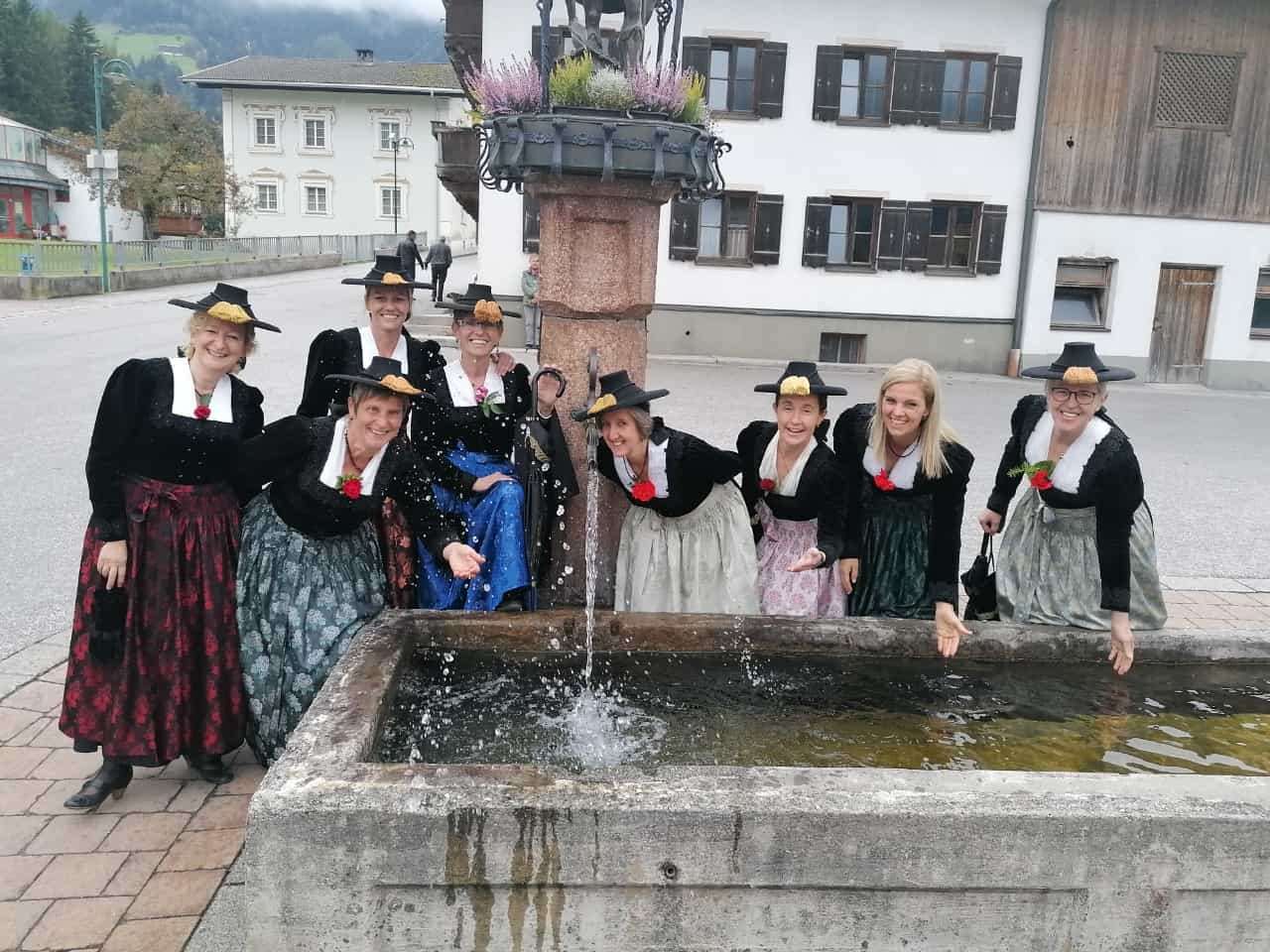 Eine Gruppe von Frauen in traditioneller Kleidung, posiert für ein Foto in der Nähe eines Brunnens. Dahinter ein Gebäude mit vielen Fenstern und einem Weg.