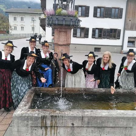 Eine Gruppe von Frauen in traditioneller Kleidung, posiert für ein Foto in der Nähe eines Brunnens. Dahinter ein Gebäude mit vielen Fenstern und einem Weg.