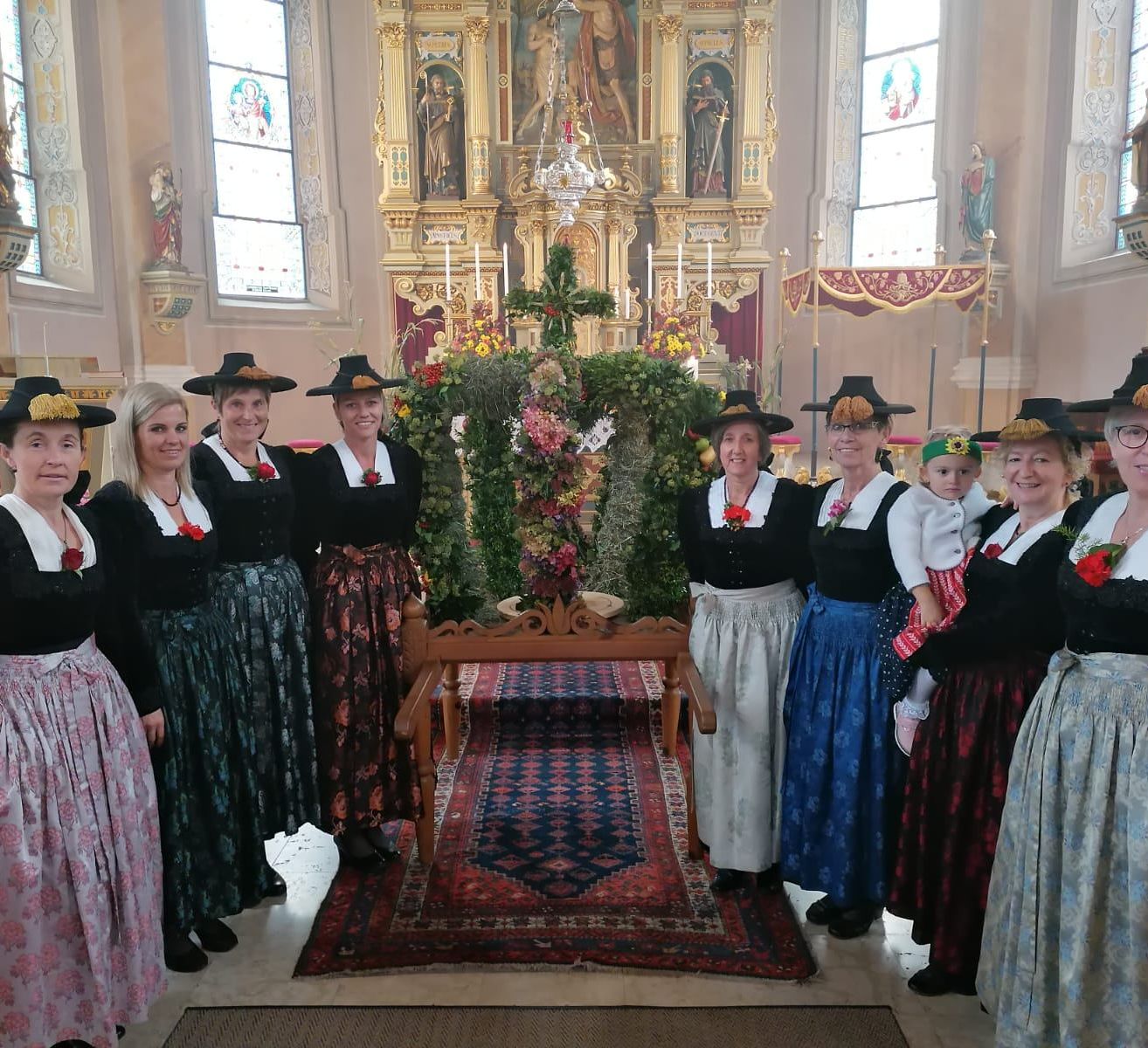 Eine Gruppe von Frauen in traditioneller Kleidung steht in einer Kirche und lächelt für ein Foto. Ein geschmückter Altar ist hinter ihnen, mit einem hölzernen Tisch und einem farbenfrohen Teppich auf dem Boden.
