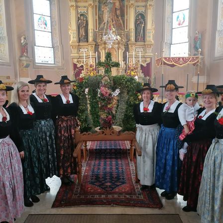 Eine Gruppe von Frauen in traditioneller Kleidung steht in einer Kirche und lächelt für ein Foto. Ein geschmückter Altar ist hinter ihnen, mit einem hölzernen Tisch und einem farbenfrohen Teppich auf dem Boden.