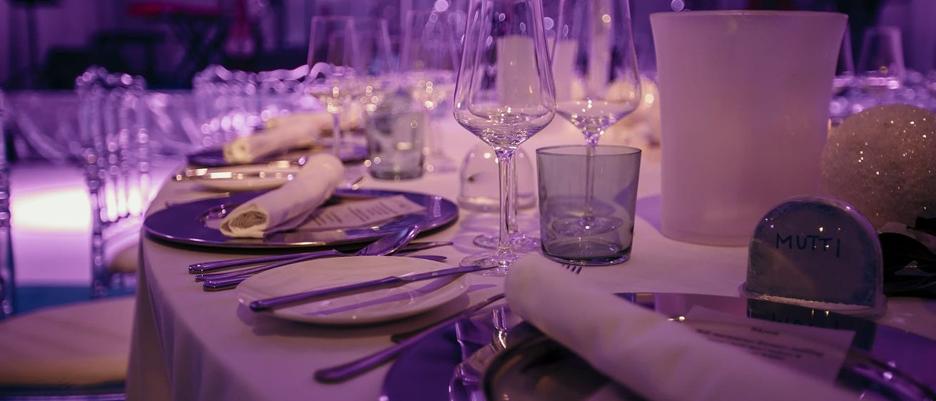 Ein Tisch mit violetter Beleuchtung, bestehend aus einer Glasvase, Weingläsern und Tellern mit Besteck und Servietten. Der Tisch ist mit violetten Tellern und weißen Geschirrstücken geschmückt.