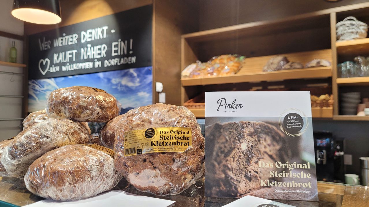 Eine Nahaufnahme von eingewickelten Broten mit dem Etikett 'Das Original Steirische Kletzenbrot', zusammen mit einem Buchcover für 'Pirker', das die gleiche Art von Brot zeigt. Dahinter zeigt ein Regal verschiedene Gebäckstücke. Ein Schild steht: 'Denkt näher ein! An Dorfladen'.