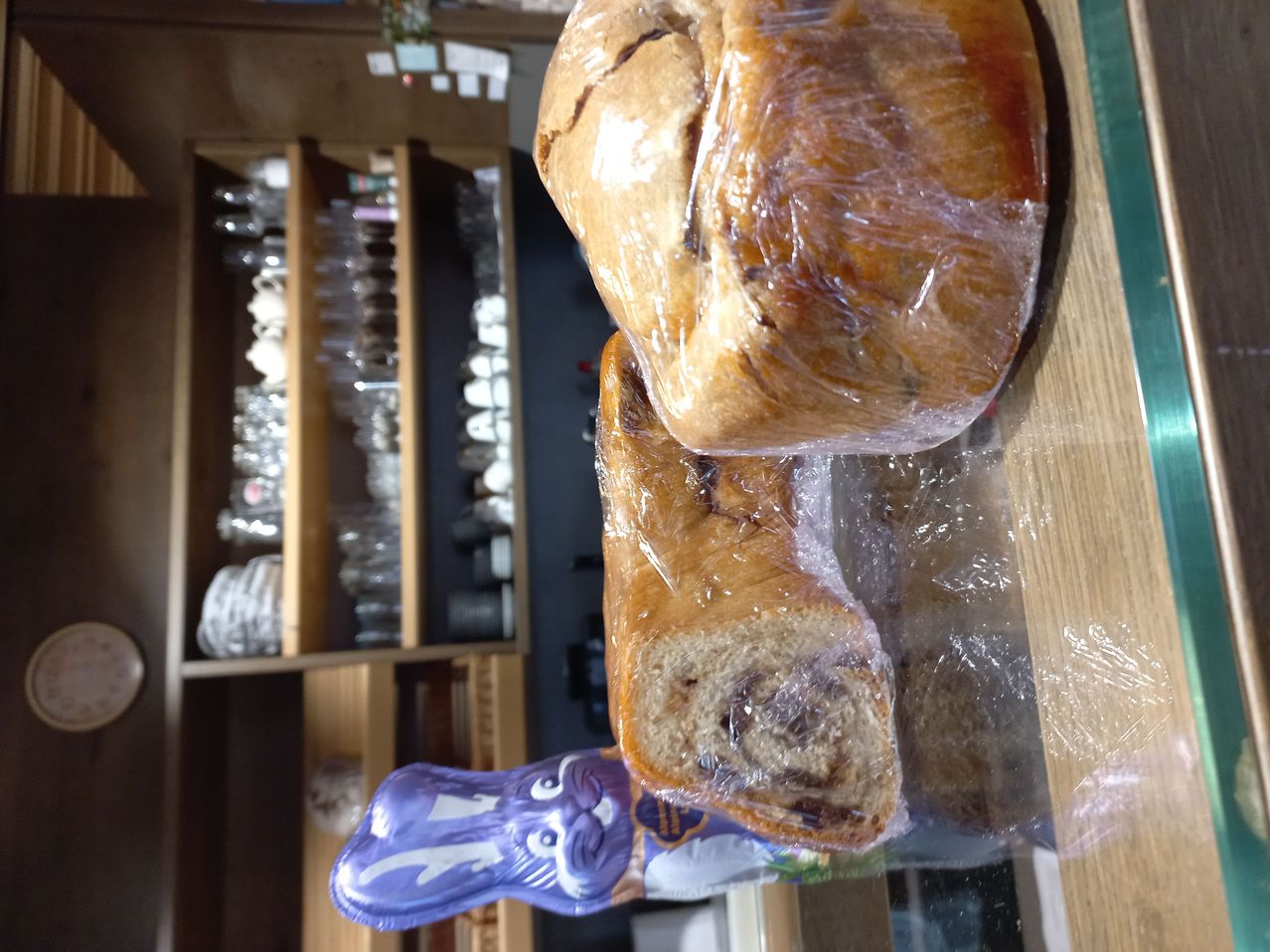 Zwei in Plastik eingewickelte Brote stehen auf einer Holzoberfläche. Dahinter befindet sich ein Holzregal mit Gläsern und Tellern.