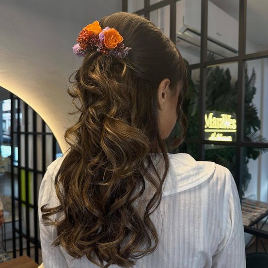 Eine Frau mit lockigem Haar und einer Blumenhaarspange steht vor einem Fenster mit einem Schild, auf dem 'Marques' steht.