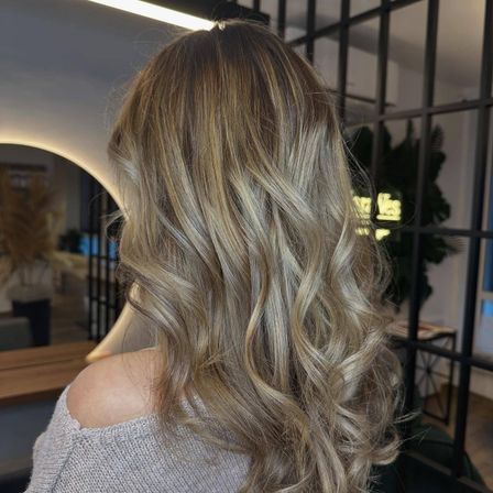 Eine Frau mit langen, welligen blonden Haaren, die in einem Salon gestylt wurde, steht vor einem Spiegel. Hinter ihr sind eine schwarze Metallstruktur und eine Pflanze zu sehen.