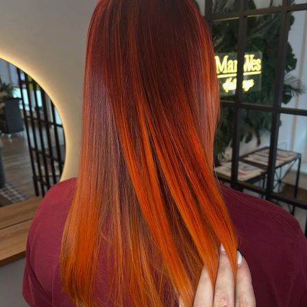 Eine Frau mit leuchtend orangefarbenen Haaren in einem Salon, mit dem Rücken zur Kamera. Im Hintergrund befindet sich ein Neonschild und eine Pflanze.