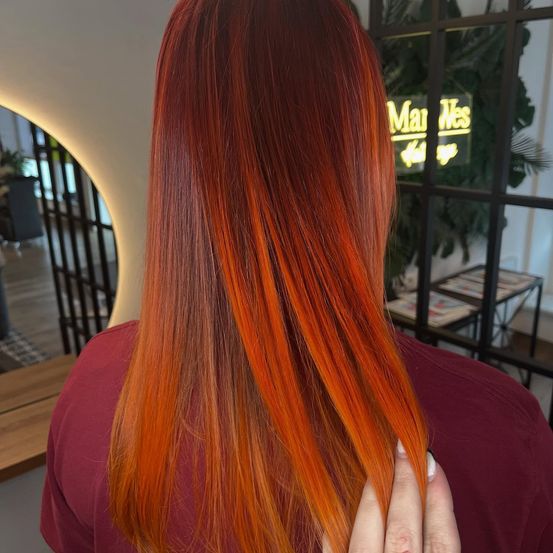 Eine Frau mit leuchtend orangefarbenen Haaren in einem Salon, mit dem Rücken zur Kamera. Im Hintergrund befindet sich ein Neonschild und eine Pflanze.