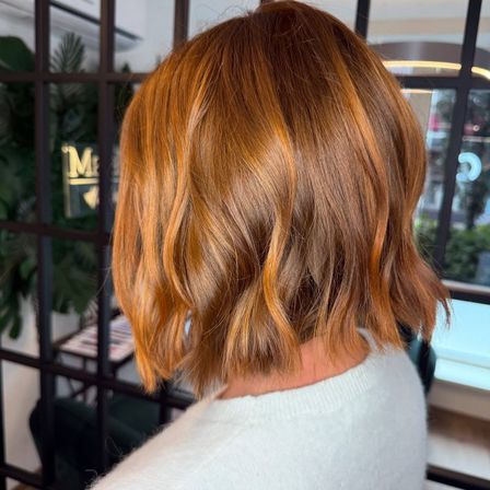 Eine Frau mit kurzen, welligen, rötlich-braunen Haaren ist von hinten zu sehen, sie trägt einen weißen Pullover. Sie steht vor einer Glaswand.