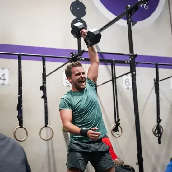 Ein Mann im Fitnessstudio hebt eine schwarze Hantel, während andere zusehen, mit nummerierten Ringen dahinter.