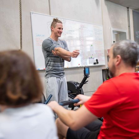 Ein Mann gibt zwei Personen auf einem Rudergerät Anweisungen, gestikuliert mit den Armen und steht vor einer Tafel. Die Umgebung scheint ein Fitnessstudio zu sein.